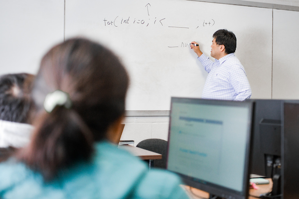 Student sitting at a computer, watching teacher write on the board