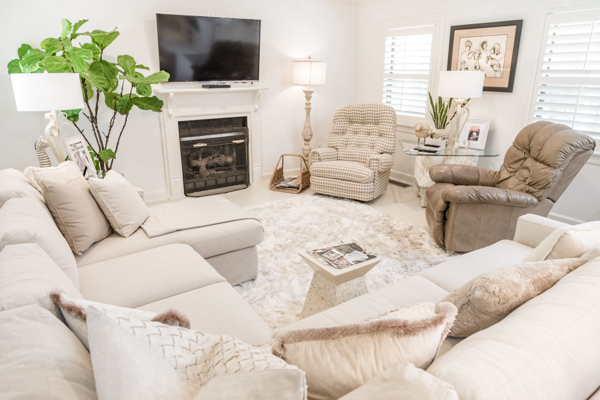 Inside the living room a white fur rug anchors the sectional couch and two armchairs in front of the fireplace that holds large screen television on top of the white mantle.