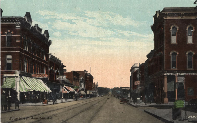 Color print of Noble Street in Anniston, Alabama showing one, two, and three-story commercial buildings and streetcar rails.