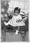 Professional portrait of Willadean Martin as ca. 6-month old child (born Aug 7 1943), child in dress, hair ribbon, socks, shoes, sitting on table, tiled floor. 